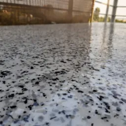 Jasnoszara, błyszcząca posadzka z czarnymi i szarymi płatkami, widok z niskiej perspektywy na zewnątrz z metalową balustradą w tle.