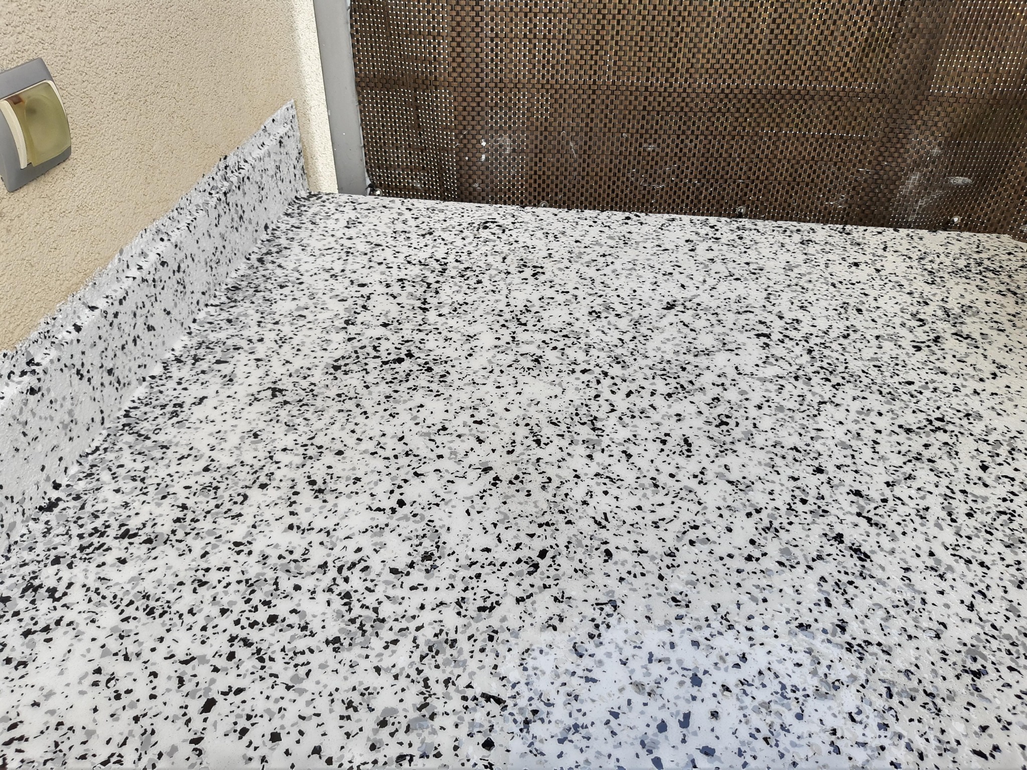 Szara i biała powierzchnia z drobnymi czarnymi plamkami, wykończona na krawędzi przy ścianie, widoczna struktura ściany i element oświetleniowy.