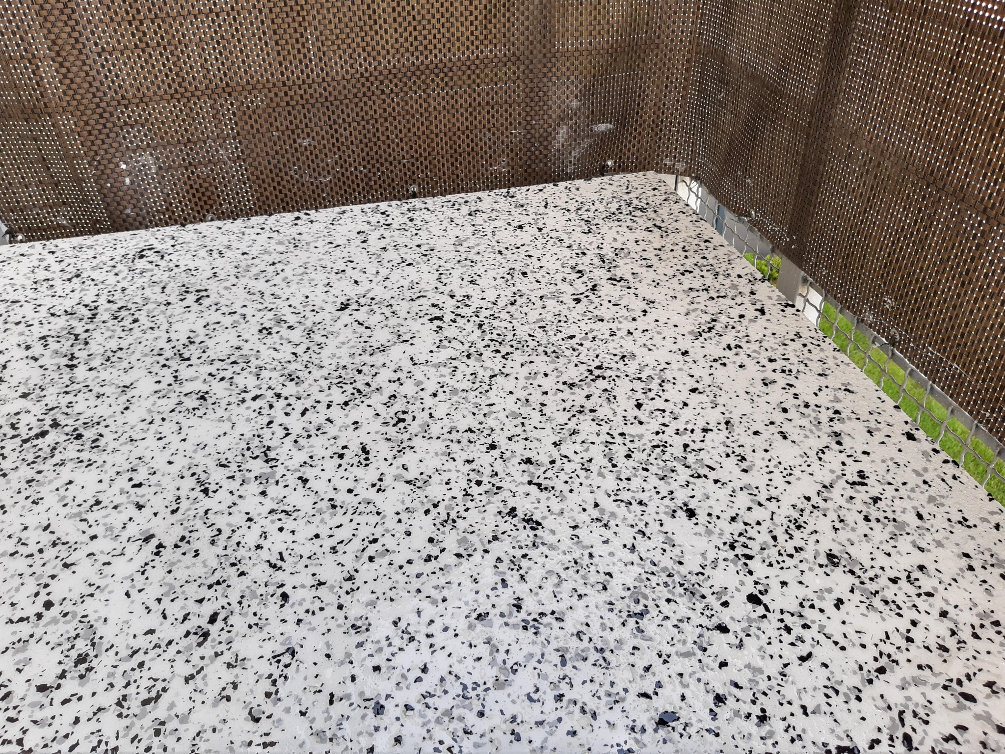 Biała powierzchnia z czarnymi i szarymi drobnymi elementami, wykończona srebrną listwą przy ścianie z dekoracyjnym panelem z brązowej plecionki, widoczny fragment zieleni.