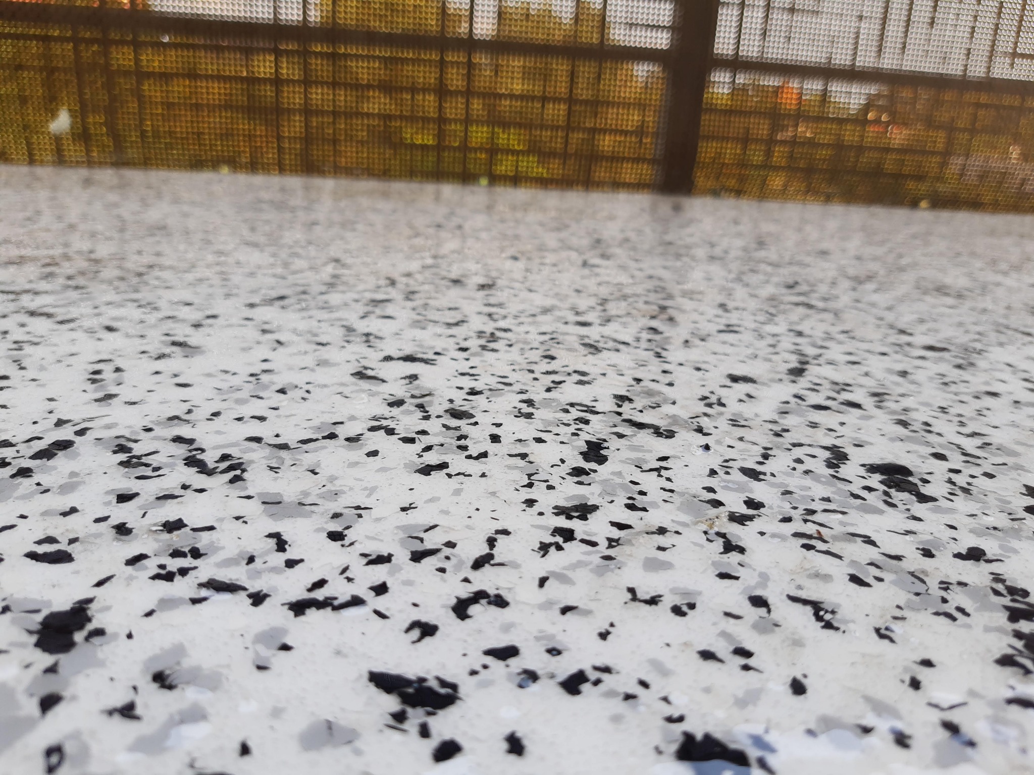 Jasna posadzka z czarnymi i szarymi płatkami, widok z niskiej perspektywy na zewnątrz, z widoczną metalową konstrukcją w tle.