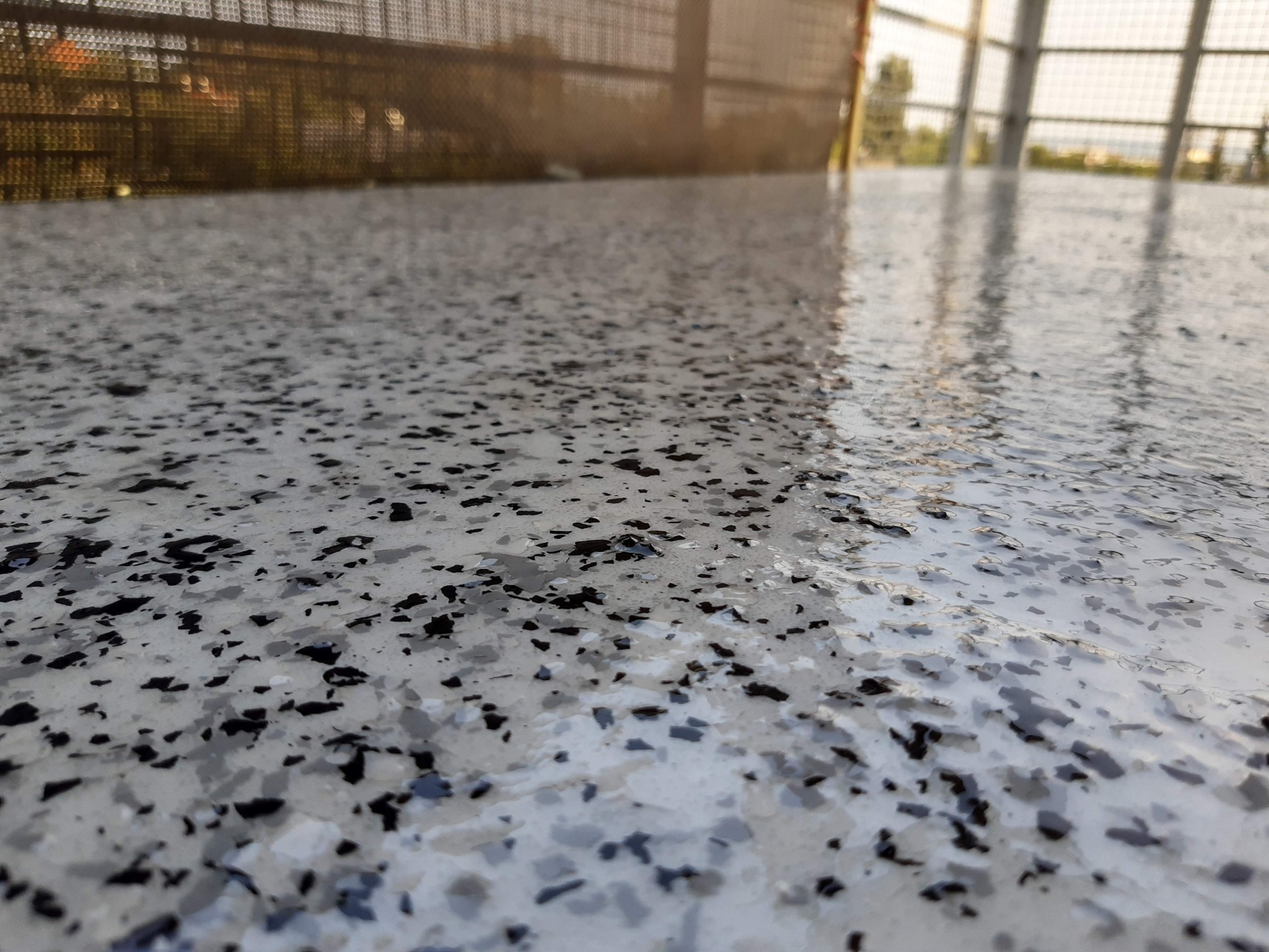 Jasnoszara, błyszcząca posadzka z czarnymi i szarymi płatkami, widok z niskiej perspektywy na zewnątrz z metalową balustradą w tle.