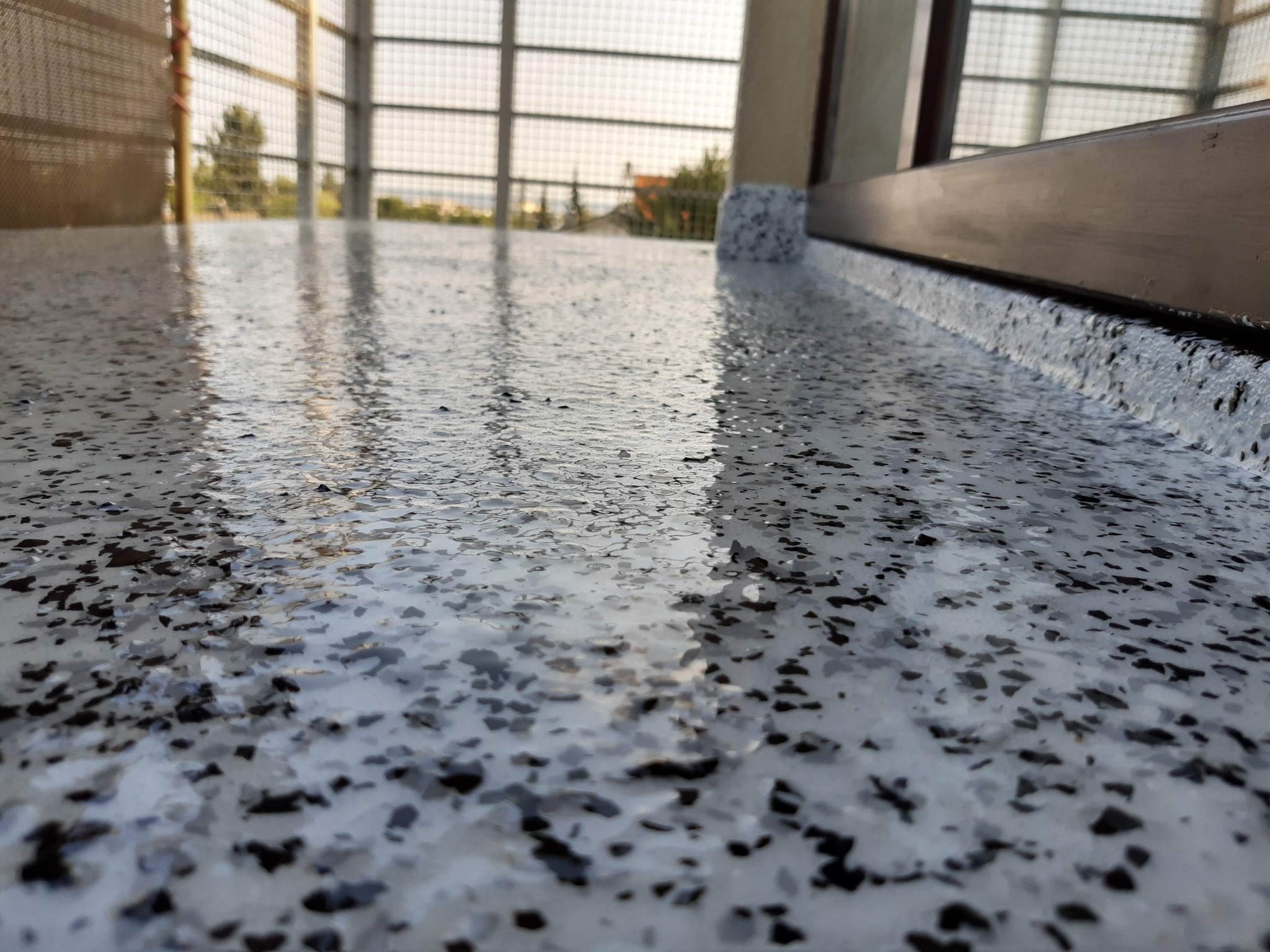 Błyszcząca, jasnoszara posadzka z drobnymi, ciemnymi wtrąceniami na balkonie, widok z niskiej perspektywy, z widoczną balustradą i fragmentem krajobrazu w tle.