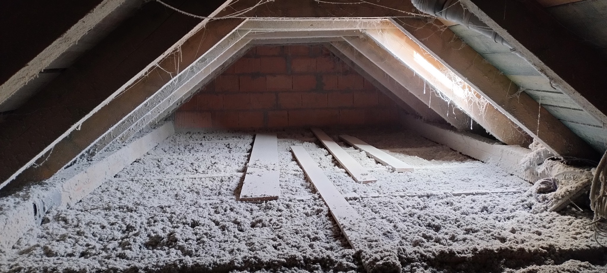 Poddasze z widoczną izolacją celulozową, drewnianą konstrukcją dachu z pajęczynami i fragmentem komina z cegły