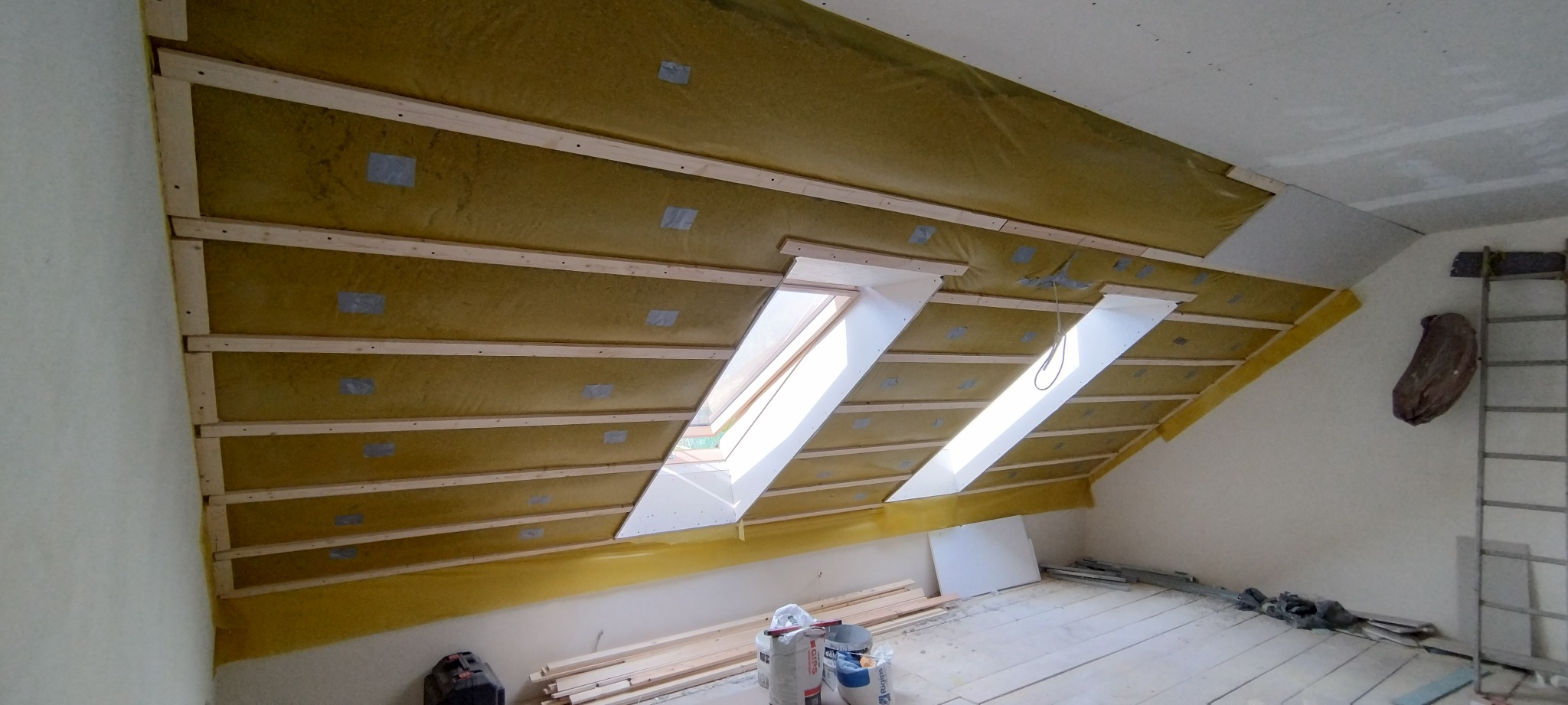 Poddasze w trakcie ocieplania wełną mineralną, widoczne drewniane legary, folia paroizolacyjna, okna dachowe, oraz drabina oparta o ścianę.