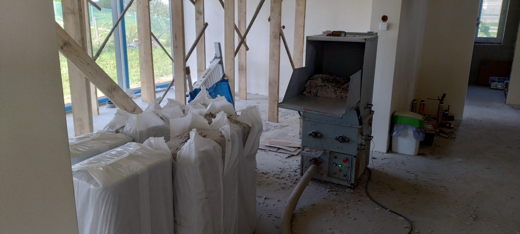 Maszyna do rozdrabniania wełny mineralnej z workami pełnymi materiału izolacyjnego w surowym wnętrzu budynku w trakcie ocieplania.