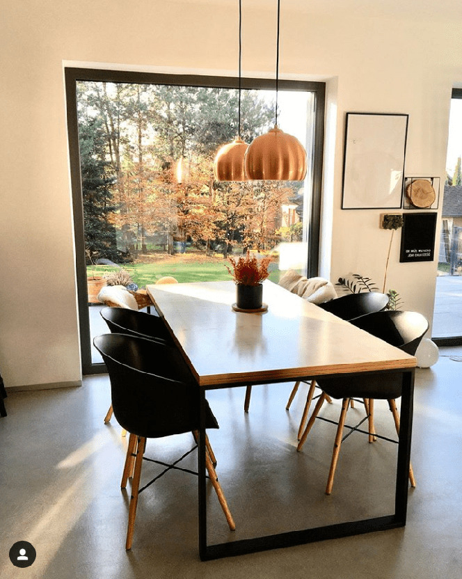 Jasna, nowoczesna jadalnia z podłogą w odcieniach szarości, stół z blatem imitującym beton, czarne krzesła z drewnianymi nogami, duże okno z widokiem na jesienny ogród, dwie miedziane lampy wiszące.