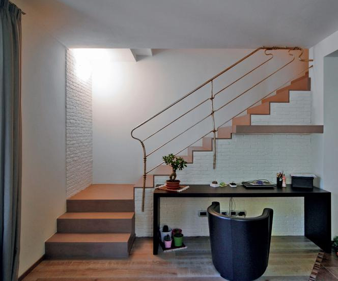 Schody z mikrocementu w nowoczesnym wnętrzu, z kontrastującym białym murem z cegły i metalową balustradą, obok biurko z bonsai.