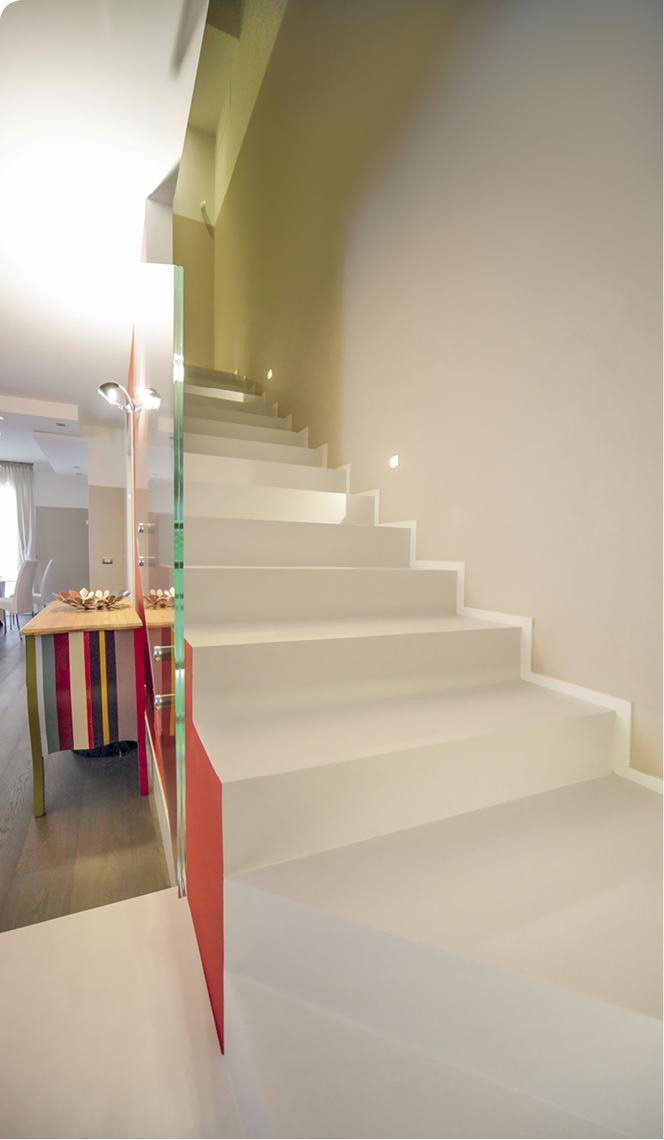 Nowoczesne, białe schody z podświetleniem LED w ścianie, zabezpieczone szklaną balustradą z czerwonym akcentem, widok na salon z kolorowym stolikiem.