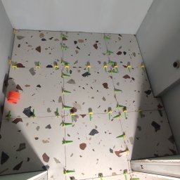 Life-bud-Mikolaj - Układanie płytek terazzo w łazience z użyciem systemu poziomowania. Widoczne zielone i żółte kliny. W tle białe ściany i fragment obuwia.