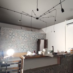 Life-bud-Mikolaj - Wnętrze kuchni w trakcie remontu: widoczne płytki na ścianie, lampy pająki na suficie, sprzęt roboczy i materiały budowlane. Etap prac wykończeniowych.
