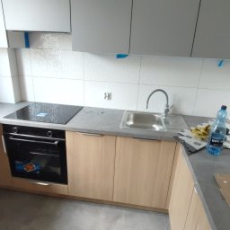 Life-bud-Mikolaj - Jasne, drewniane szafki kuchenne z szarym blatem i płytą indukcyjną. Białe, heksagonalne płytki na ścianie. Nowoczesna armatura i butelka wody. Podłoga wyłożona szarymi płytkami.