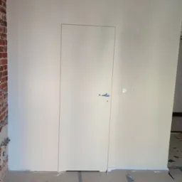 Białe, minimalistyczne drzwi ukryte w ścianie, z widocznymi włącznikami i fragmentem ceglanej ściany. Podłoga zabezpieczona folią malarską.