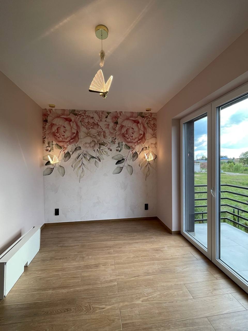 Pokój z drewnopodobną podłogą, ścianą w kwiaty i motylkową lampą. Okno balkonowe z widokiem na zewnątrz. Minimalistyczny grzejnik pod ścianą. Nowoczesne wykończenie.