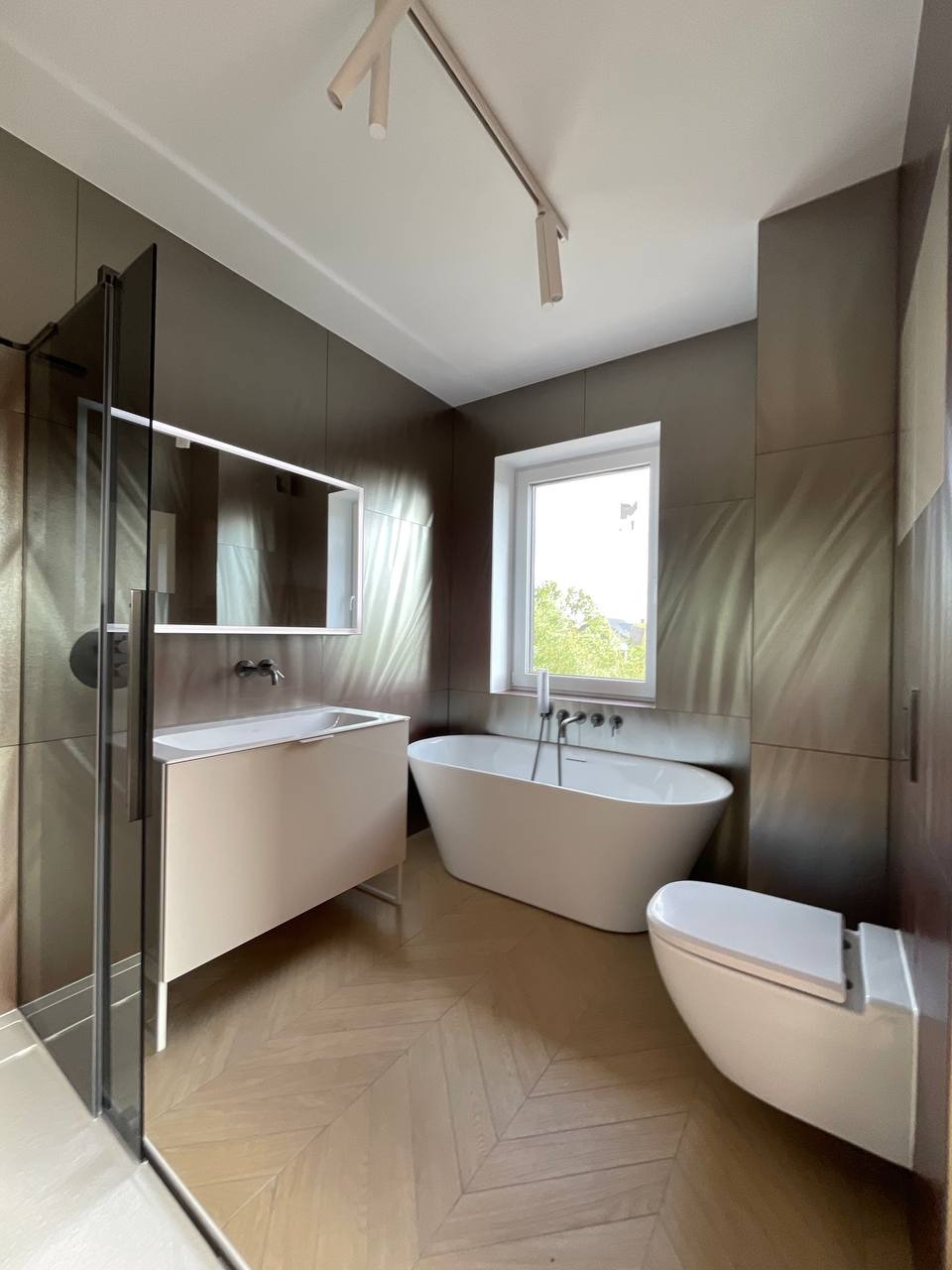 Nowoczesna łazienka z kabiną prysznicową, wolnostojącą wanną i umywalką na tle płytek imitujących stal. Parkiet ułożony w jodełkę, oświetlenie szynowe.