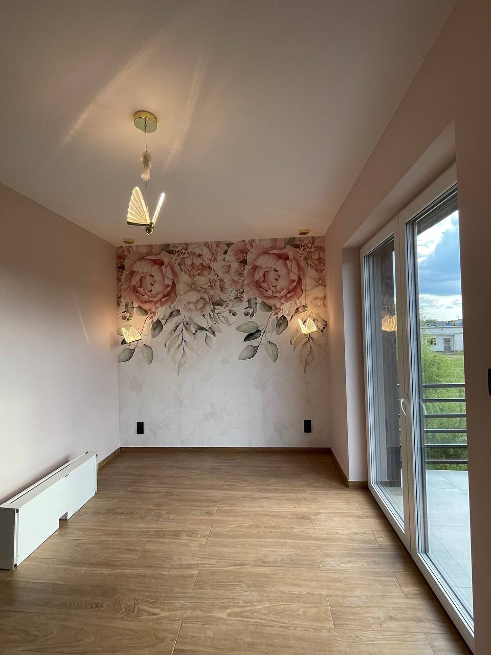 Jasne wnętrze po remoncie: drewnopodobna podłoga, ściana z motywem kwiatowym, nowoczesne lampy w kształcie motyli i duże okno balkonowe.