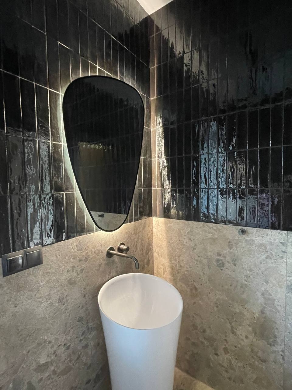 Nowoczesna łazienka z białą, wolnostojącą umywalką i czarnymi, błyszczącymi płytkami ułożonymi pionowo. Asymetryczne lustro z podświetleniem LED.