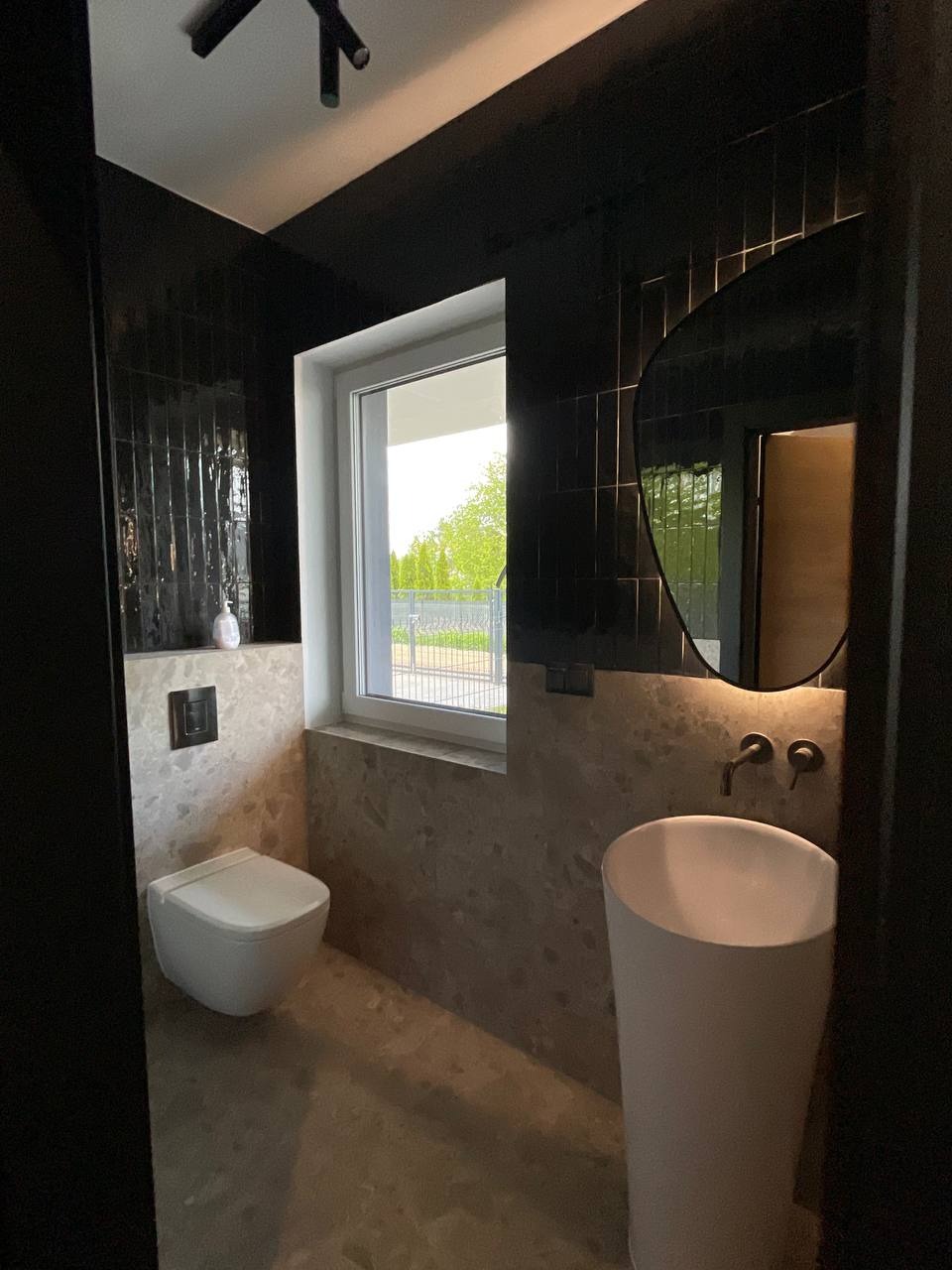 Nowoczesna łazienka z czarnymi płytkami na ścianach, owalnym lustrem i wolnostojącą umywalką. Beżowe płytki na podłodze i ścianie za toaletą. Minimalistyczne oświetlenie.