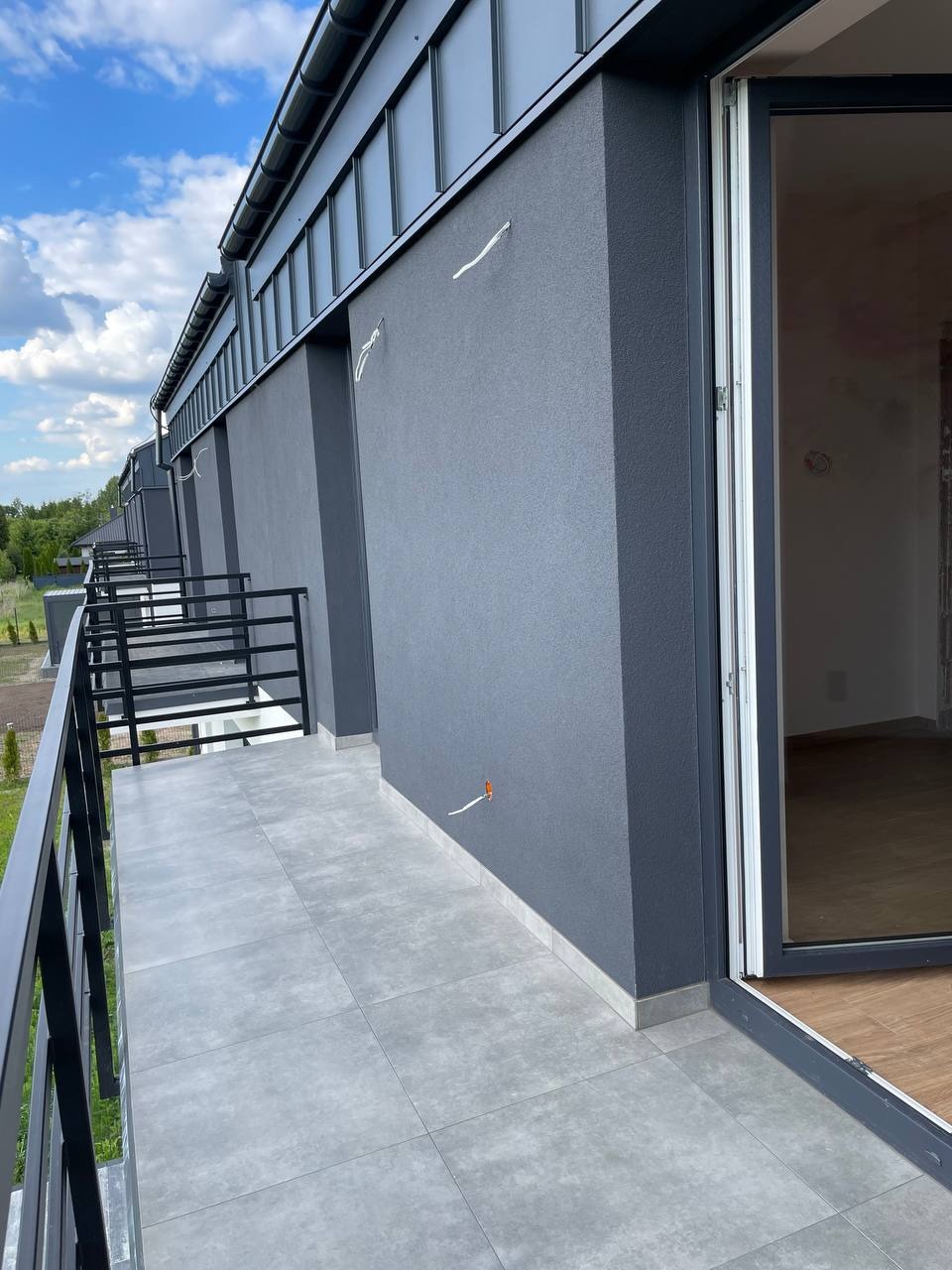 Szare płytki na balkonie nowego budynku z szarą elewacją. Widoczne wystające kable i fragment wnętrza z drewnianą podłogą. Ujęcie z dołu, niebo z chmurami w tle.