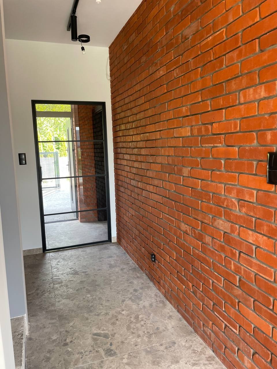 Korytarz z ceglaną ścianą w odcieniach czerwieni i szarości, z czarną ramą drzwiową i szarą podłogą. Widoczne oświetlenie punktowe na suficie i fragment otoczenia za drzwiami.