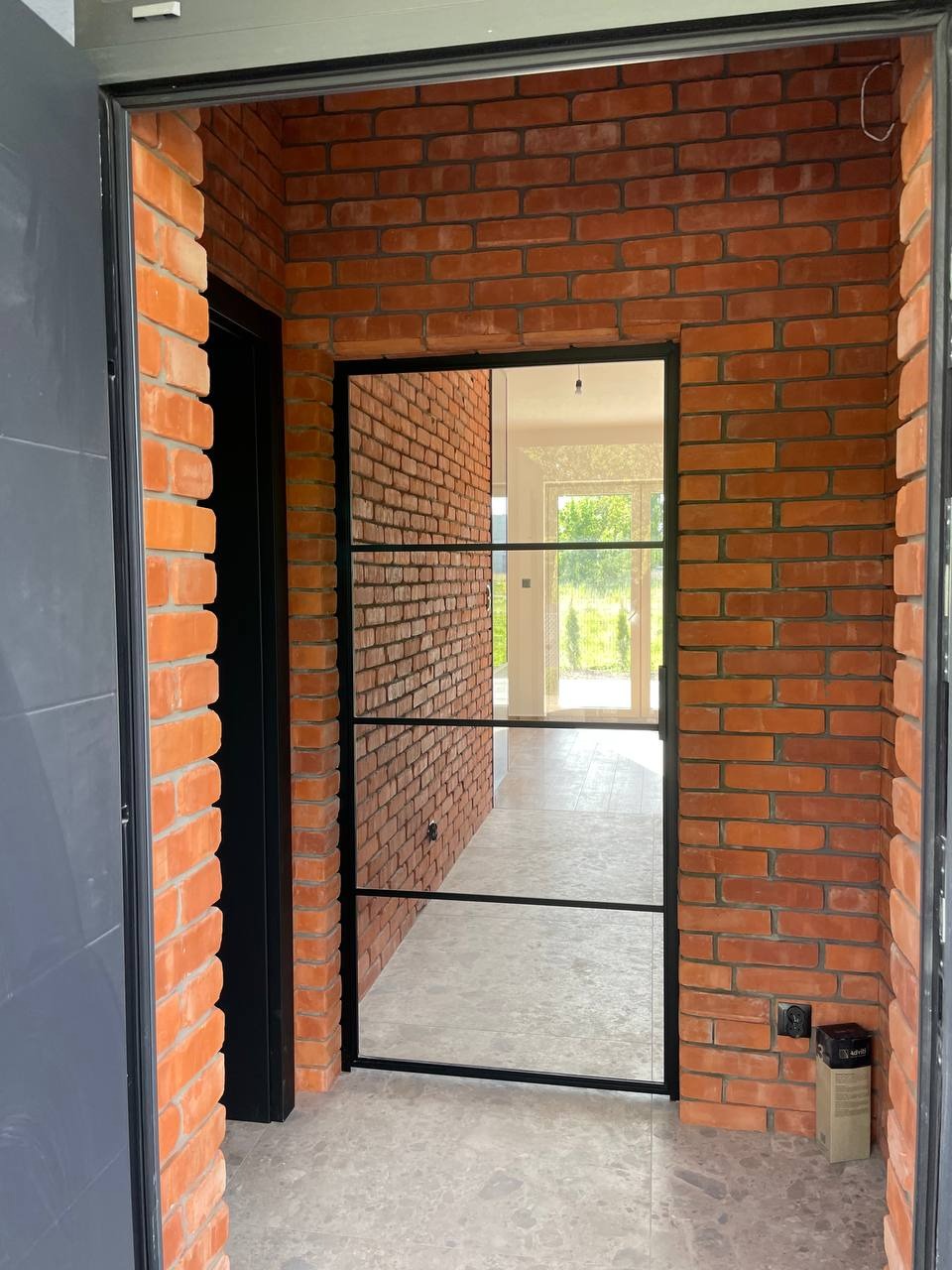 Korytarz z ceglaną ścianą i szklanymi drzwiami z czarną ramą. Widok na jasne wnętrze domu z zielenią za oknem. Nowoczesne wykończenie wnętrza.