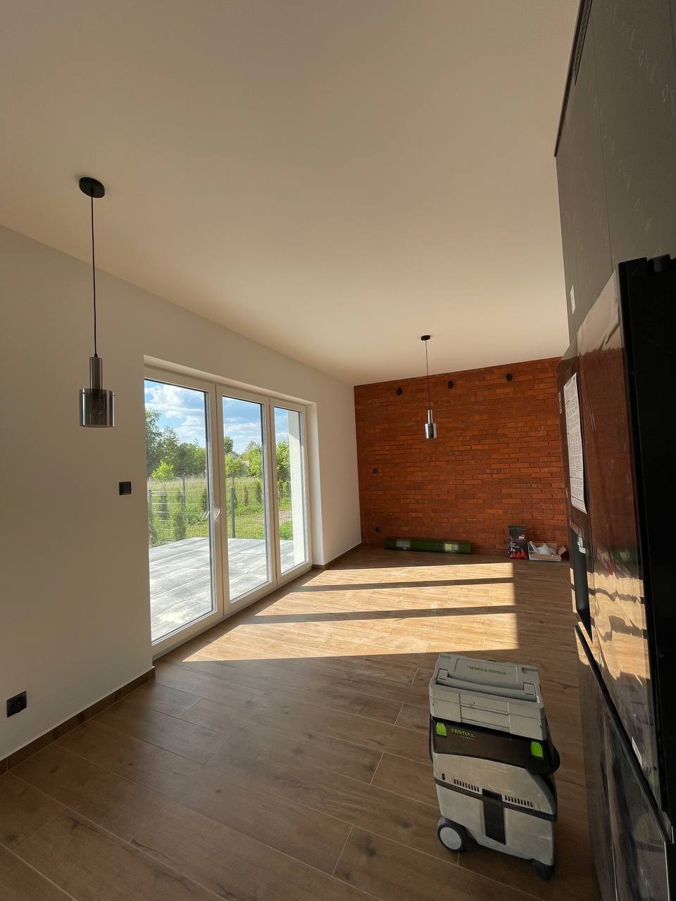 Jasne wnętrze z panelami podłogowymi, ceglaną ścianą i dużymi oknami. Widoczne narzędzia sugerują prace wykończeniowe w toku.