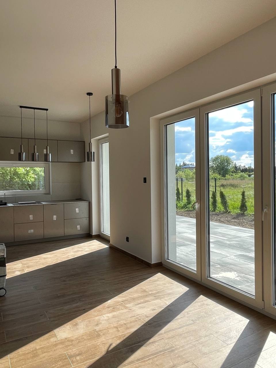 Jasne wnętrze po remoncie w Łodzi: brązowe płytki podłogowe, nowoczesne lampy wiszące, okna z widokiem na zewnątrz. Kuchnia z szarymi szafkami w tle.