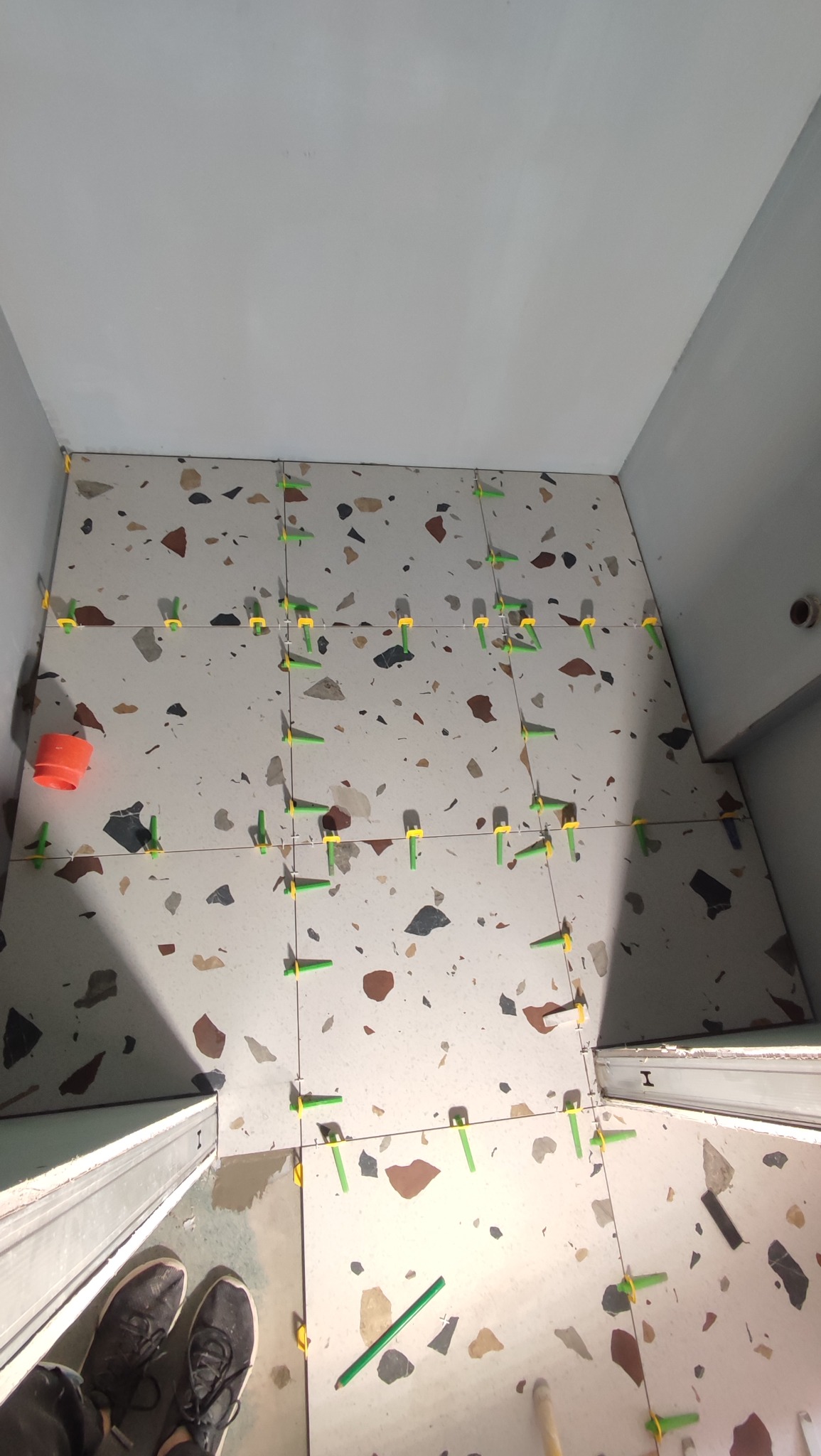 Układanie płytek terazzo w łazience z użyciem systemu poziomowania. Widoczne zielone i żółte kliny. W tle białe ściany i fragment obuwia.