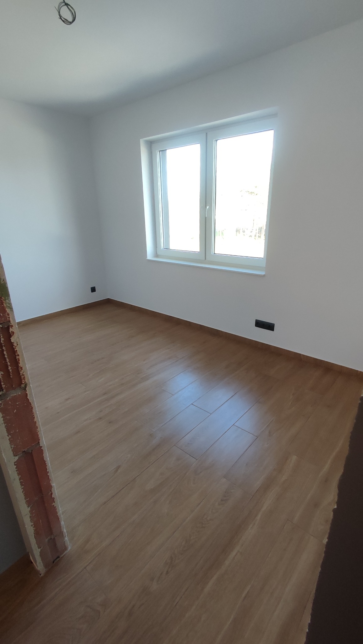 Jasne panele podłogowe w pokoju po remoncie, białe ściany i okno. Widoczny fragment muru w tle. Wnętrze po wykończeniu, gotowe do umeblowania.