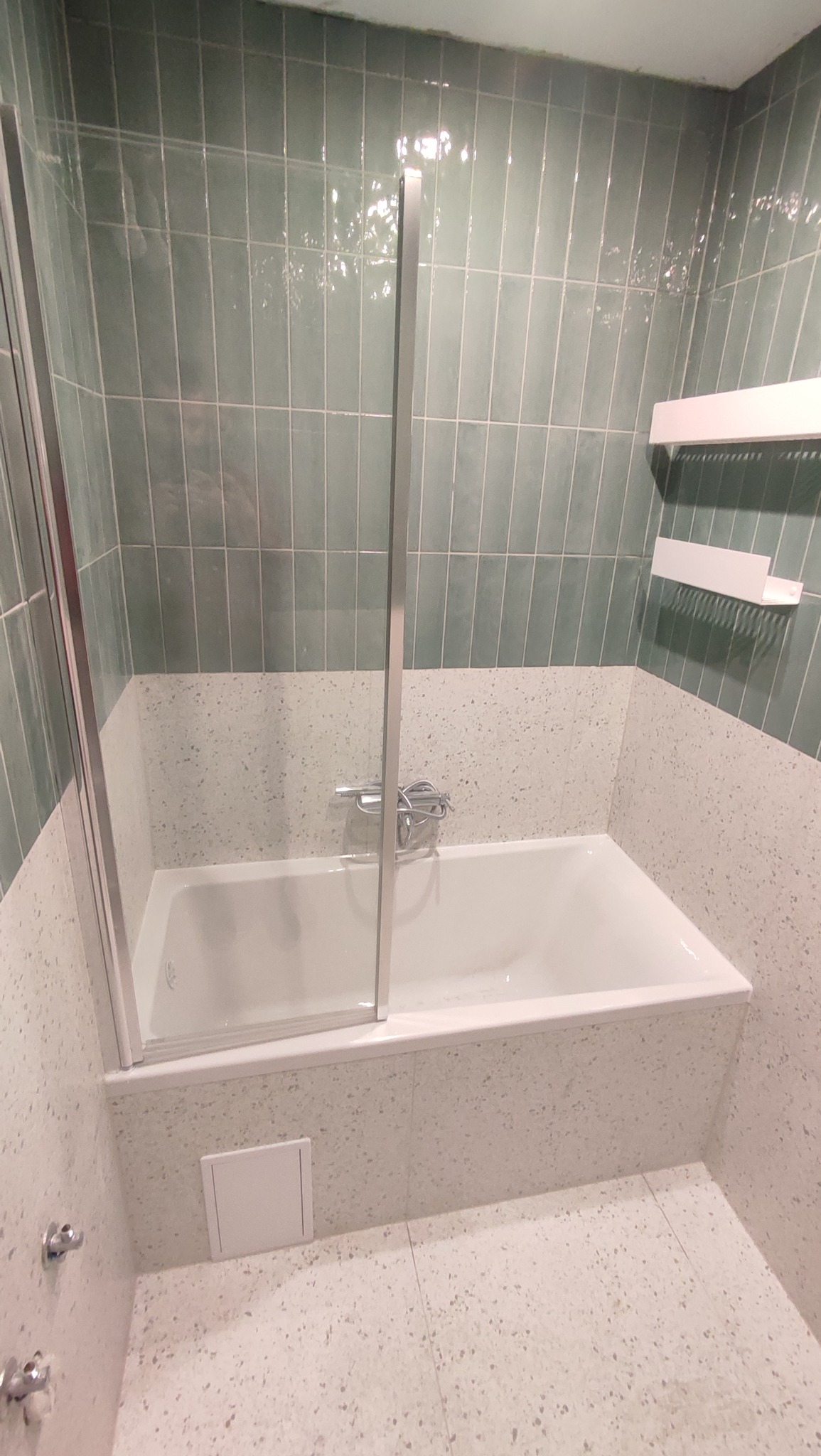 Wykończona łazienka z wanną, szklanym parawanem i zielonymi, pionowo ułożonymi płytkami. Ściany i podłoga wykończone drobnym lastryko. Minimalistyczne półki.