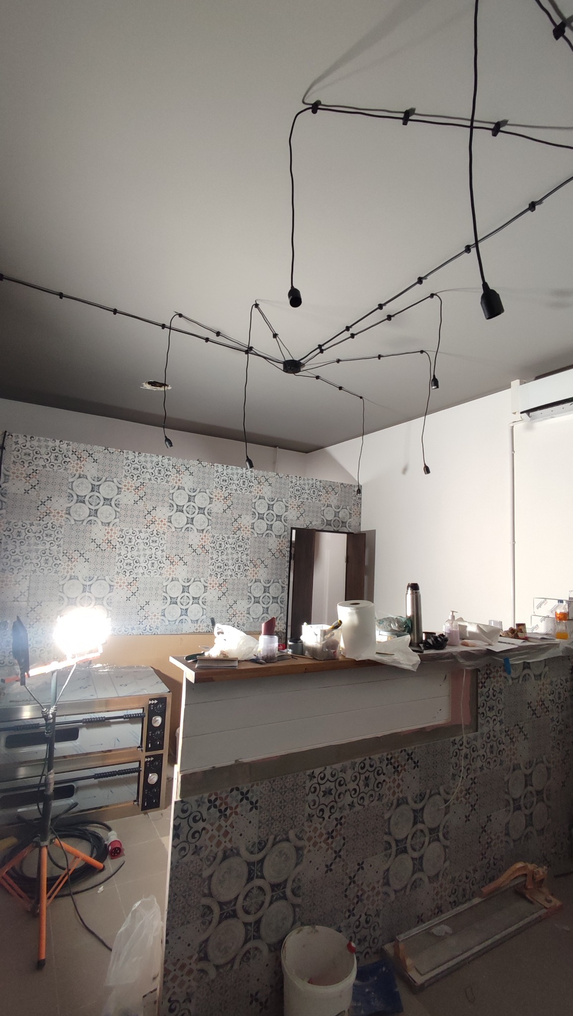 Wnętrze kuchni w trakcie remontu: widoczne płytki na ścianie, lampy pająki na suficie, sprzęt roboczy i materiały budowlane. Etap prac wykończeniowych.