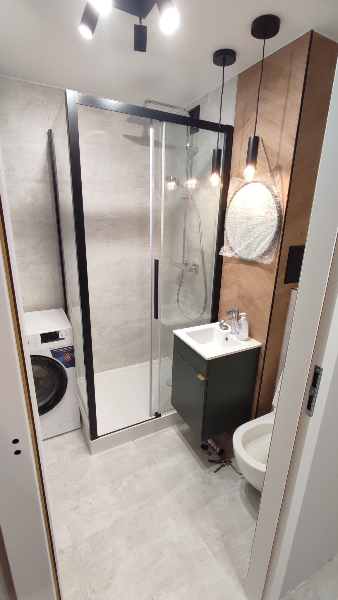 Nowoczesna łazienka po remoncie: kabina prysznicowa z czarnymi ramami, umywalka z szafką, toaleta i pralka. Oświetlenie punktowe i dekoracyjne lampy wiszące.
