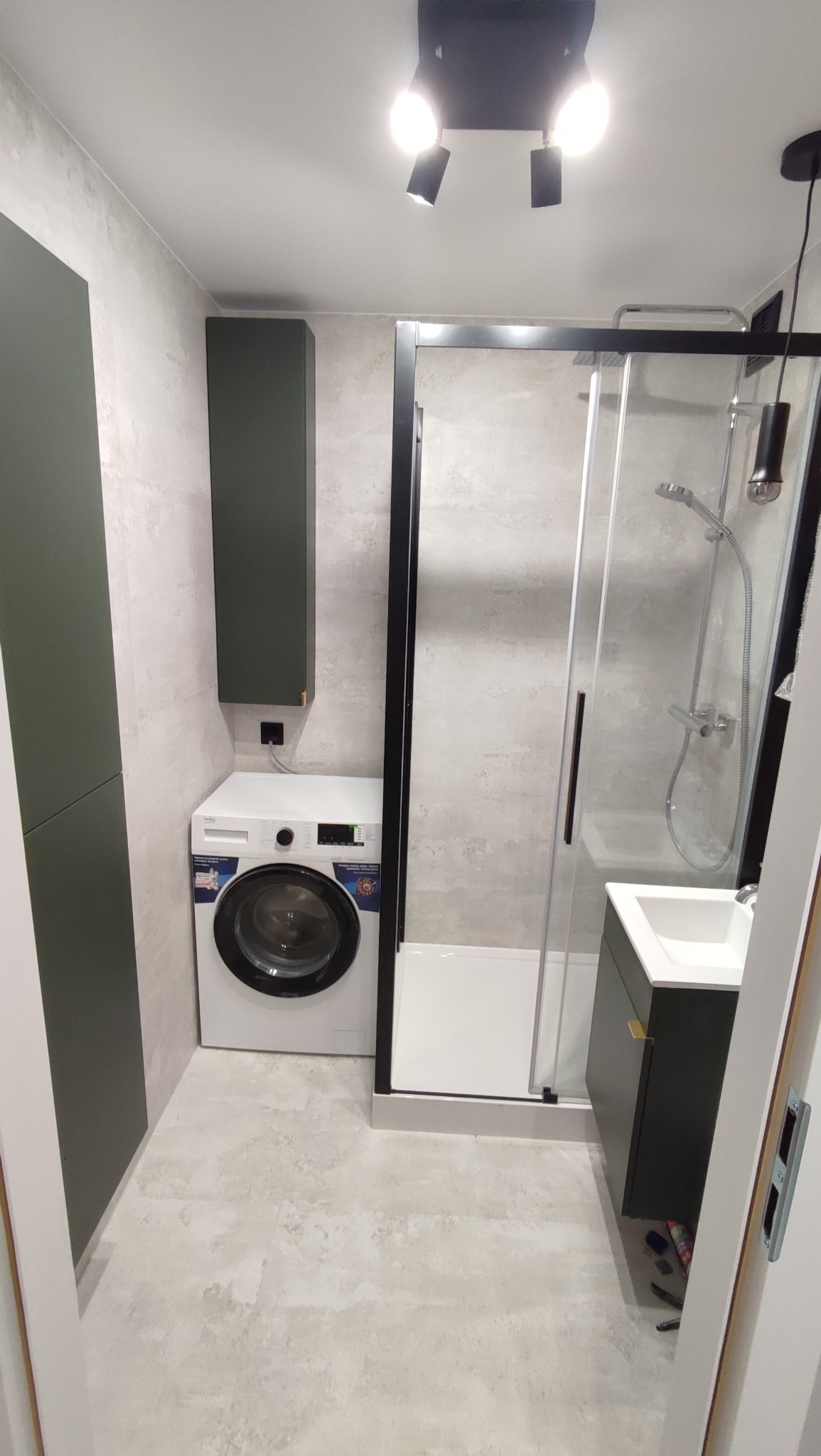 Nowoczesna łazienka z kabiną prysznicową typu walk-in, pralką i szafkami w odcieniach szarości i zieleni. Minimalistyczne oświetlenie sufitowe.