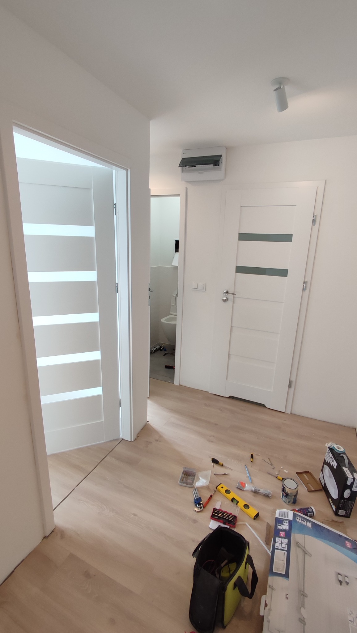 Białe drzwi z poziomymi przeszkleniami w jasnym wnętrzu, narzędzia na podłodze. Widok na białą łazienkę w tle. Minimalistyczny design.