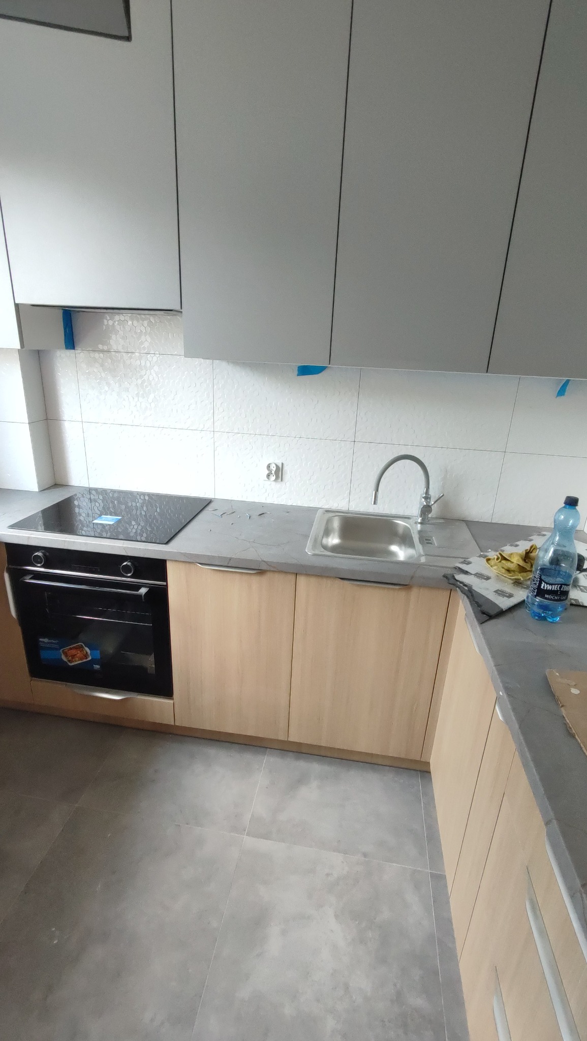 Jasne, drewniane szafki kuchenne z szarym blatem i płytą indukcyjną. Białe, heksagonalne płytki na ścianie. Nowoczesna armatura i butelka wody. Podłoga wyłożona szarymi płytkami.