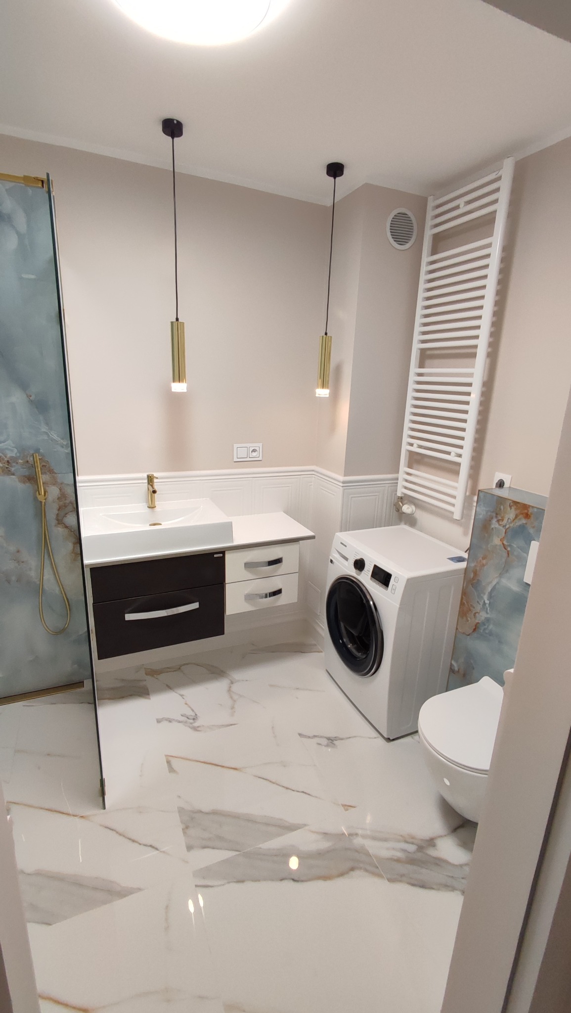 Nowoczesna łazienka w beżach z marmurową podłogą, kabiną prysznicową, umywalką z szafką, pralką i grzejnikiem drabinkowym. Dwie złote lampy wiszące.