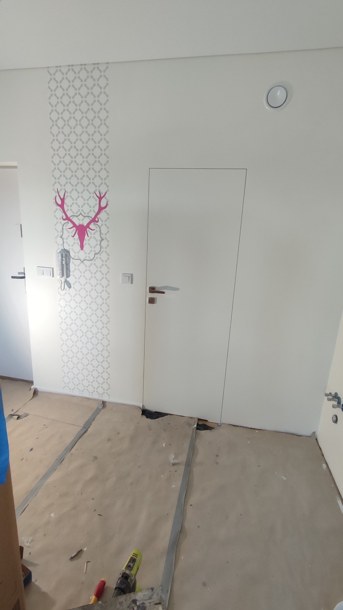 Biała ściana z szarym wzorem i różowym motywem jelenia nad domofonem, obok ukryte drzwi. Podłoga zabezpieczona kartonem i narzędzia.
