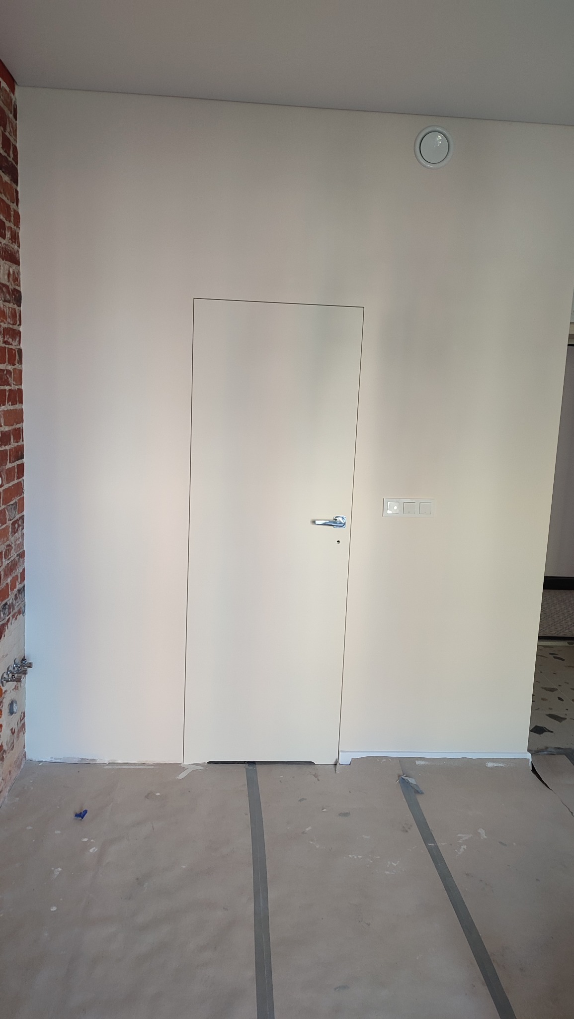 Białe, minimalistyczne drzwi ukryte w ścianie, z widocznymi włącznikami i fragmentem ceglanej ściany. Podłoga zabezpieczona folią malarską.