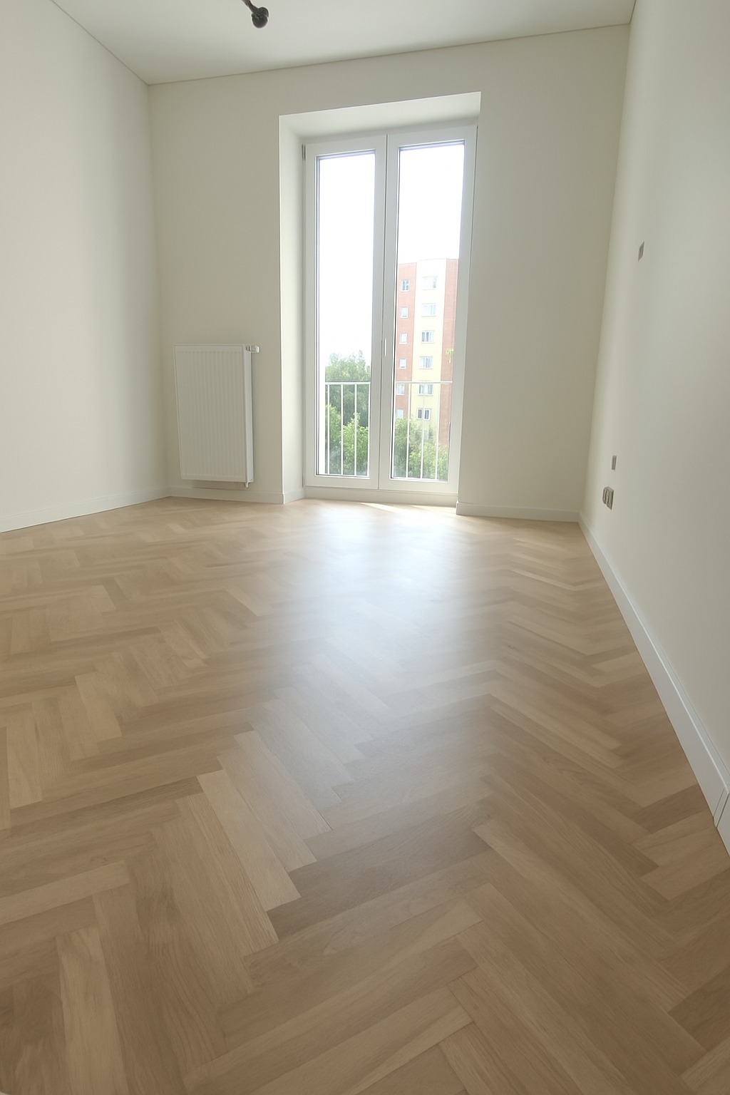 Jasne panele ułożone w jodełkę w nowym pokoju w Łodzi. Białe ściany, okno z widokiem na blok. Minimalistyczny grzejnik. Listwy przypodłogowe.