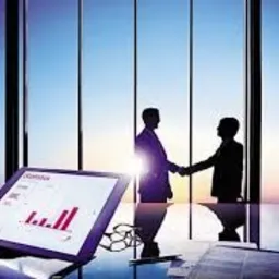 Silhouettes dwóch mężczyzn w garniturach ściskających sobie dłonie na tle okien wieżowca z widokiem na miasto o wschodzie słońca, z tabletem wyświetlającym wykresy na pierwszym planie.