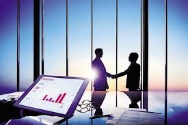 Silhouettes dwóch mężczyzn w garniturach ściskających sobie dłonie na tle okien wieżowca z widokiem na miasto o wschodzie słońca, z tabletem wyświetlającym wykresy na pierwszym planie.