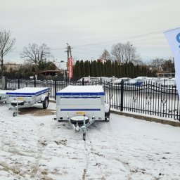 OKwynajem.pl WYPOŻYCZALNIA PRZYCZEP Radosław Witkowski - Plac z trzema przyczepami samochodowymi marki Wiola, przykrytymi plandekami w zimowy dzień, widoczna flaga reklamowa i ogrodzenie.