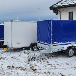 OKwynajem.pl WYPOŻYCZALNIA PRZYCZEP Radosław Witkowski - Trzy przyczepy różnych typów (plandeka, zamknięta) zaparkowane na śniegu przed budynkiem w pochmurny dzień.