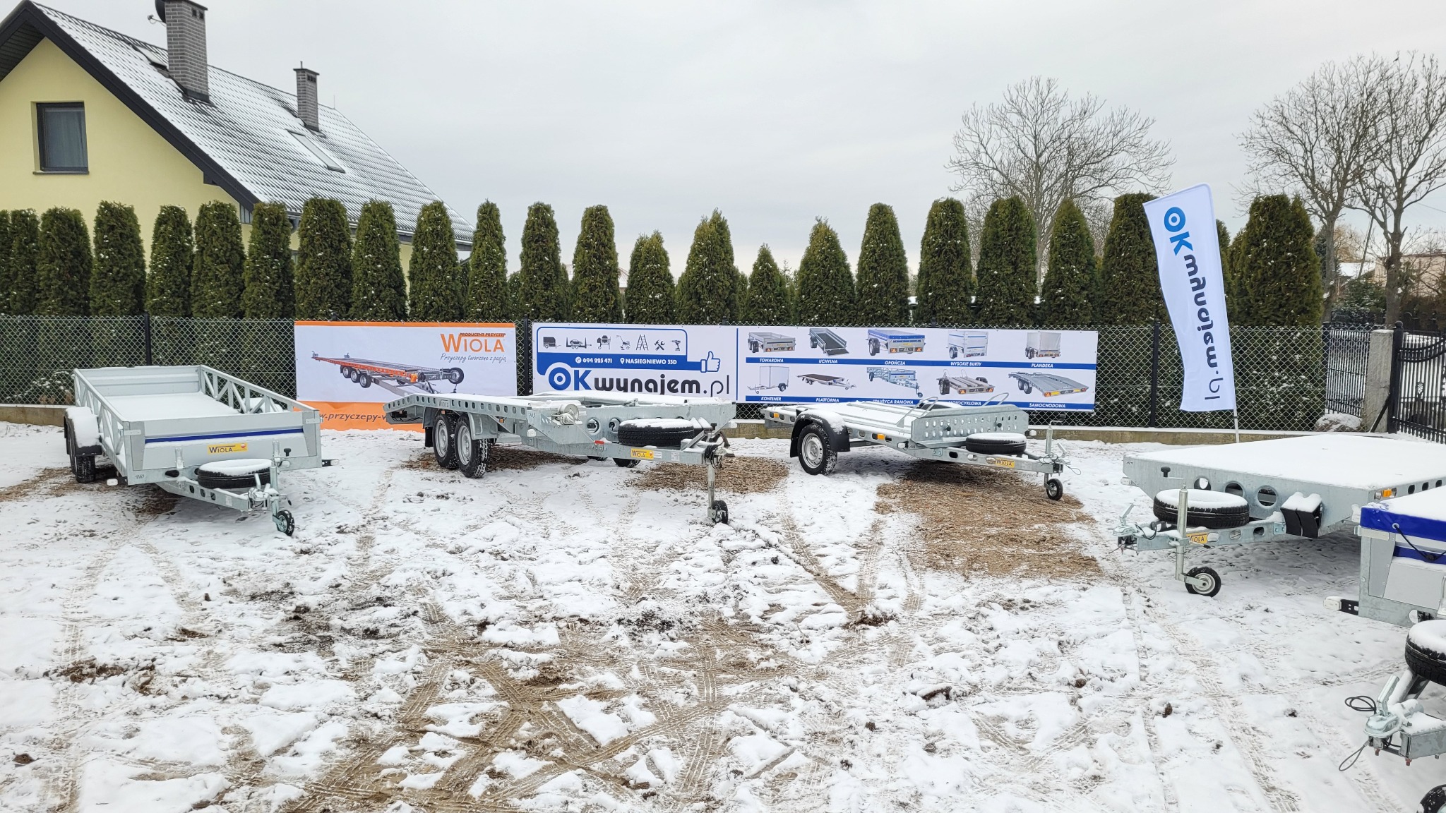 Prezentacja różnych modeli przyczep samochodowych na placu pokrytym śniegiem, z banerami reklamowymi producenta w tle i flagą firmy po prawej stronie.