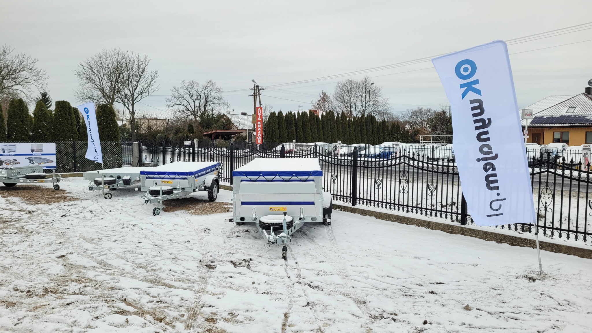 Plac z trzema przyczepami samochodowymi marki Wiola, przykrytymi plandekami w zimowy dzień, widoczna flaga reklamowa i ogrodzenie.