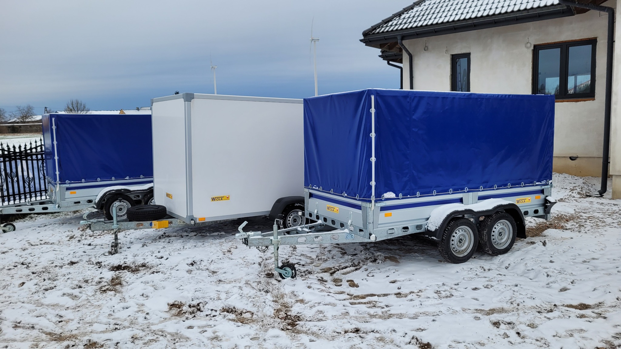 Trzy przyczepy różnych typów (plandeka, zamknięta) zaparkowane na śniegu przed budynkiem w pochmurny dzień.