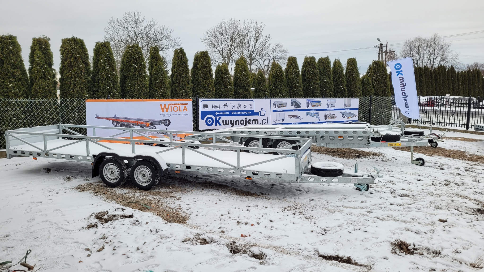 Dwie nowe, puste przyczepy samochodowe marki Wiola stojące na śniegu, z reklamami producenta w tle i flagą z logo OKwynajem.pl