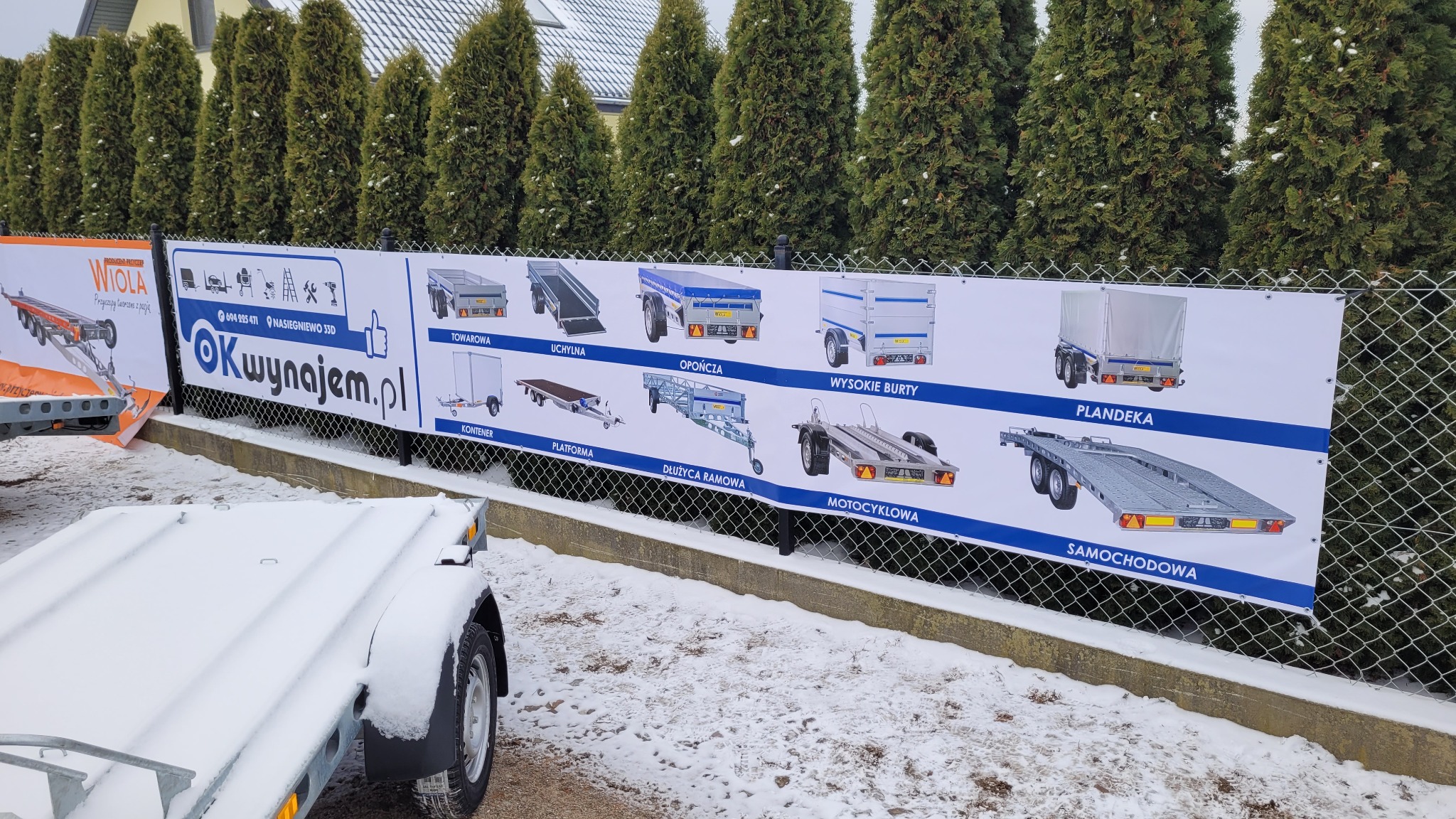Baner reklamowy z ofertą różnych typów przyczep na ogrodzeniu z siatki, częściowo zasypane śniegiem, widok z perspektywy niskiego kąta.