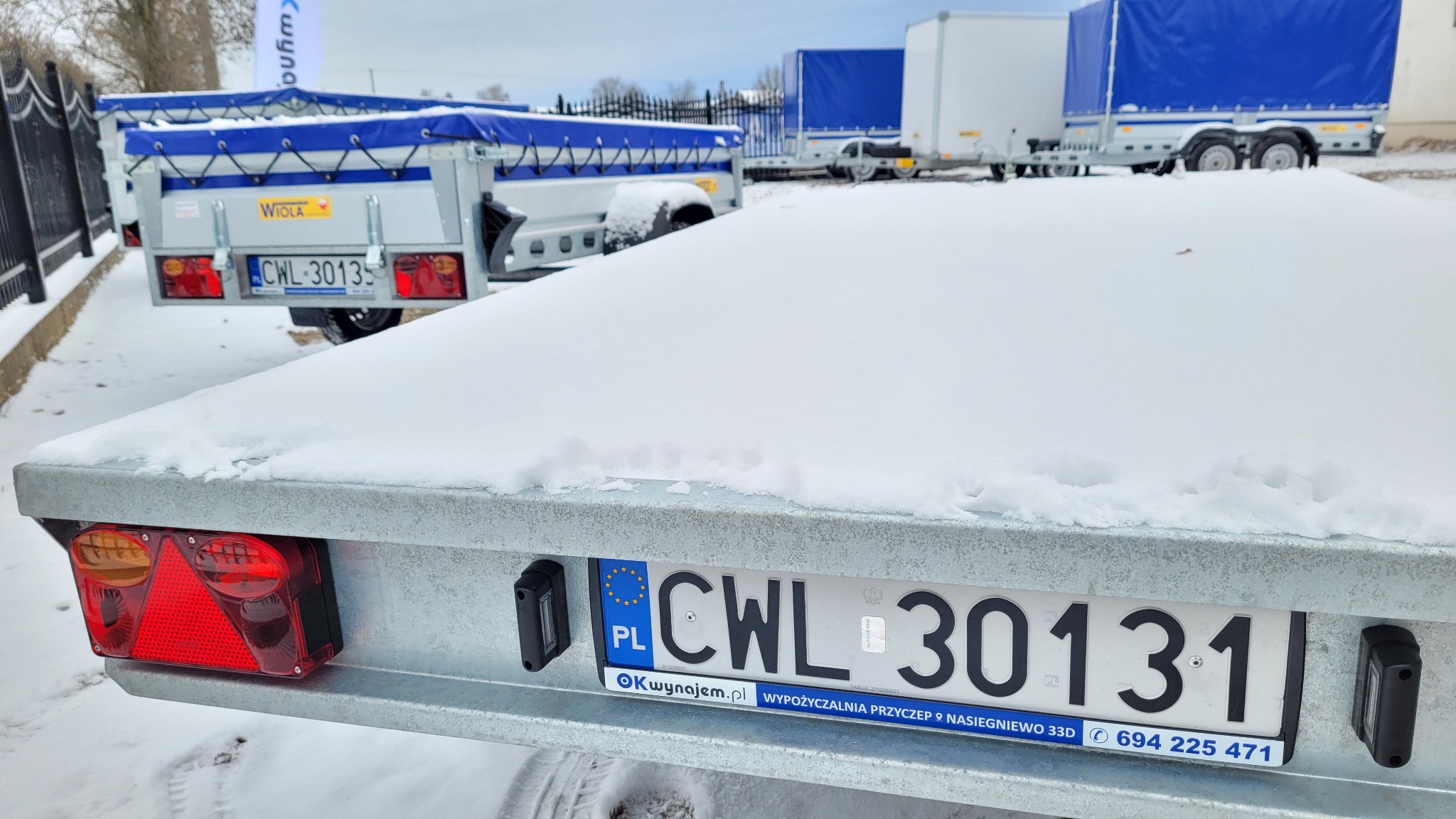 Przyczepa samochodowa pokryta śniegiem, widoczny fragment z tablicą rejestracyjną CWL 30131 i adresem wypożyczalni przyczep na tle innych przyczep.