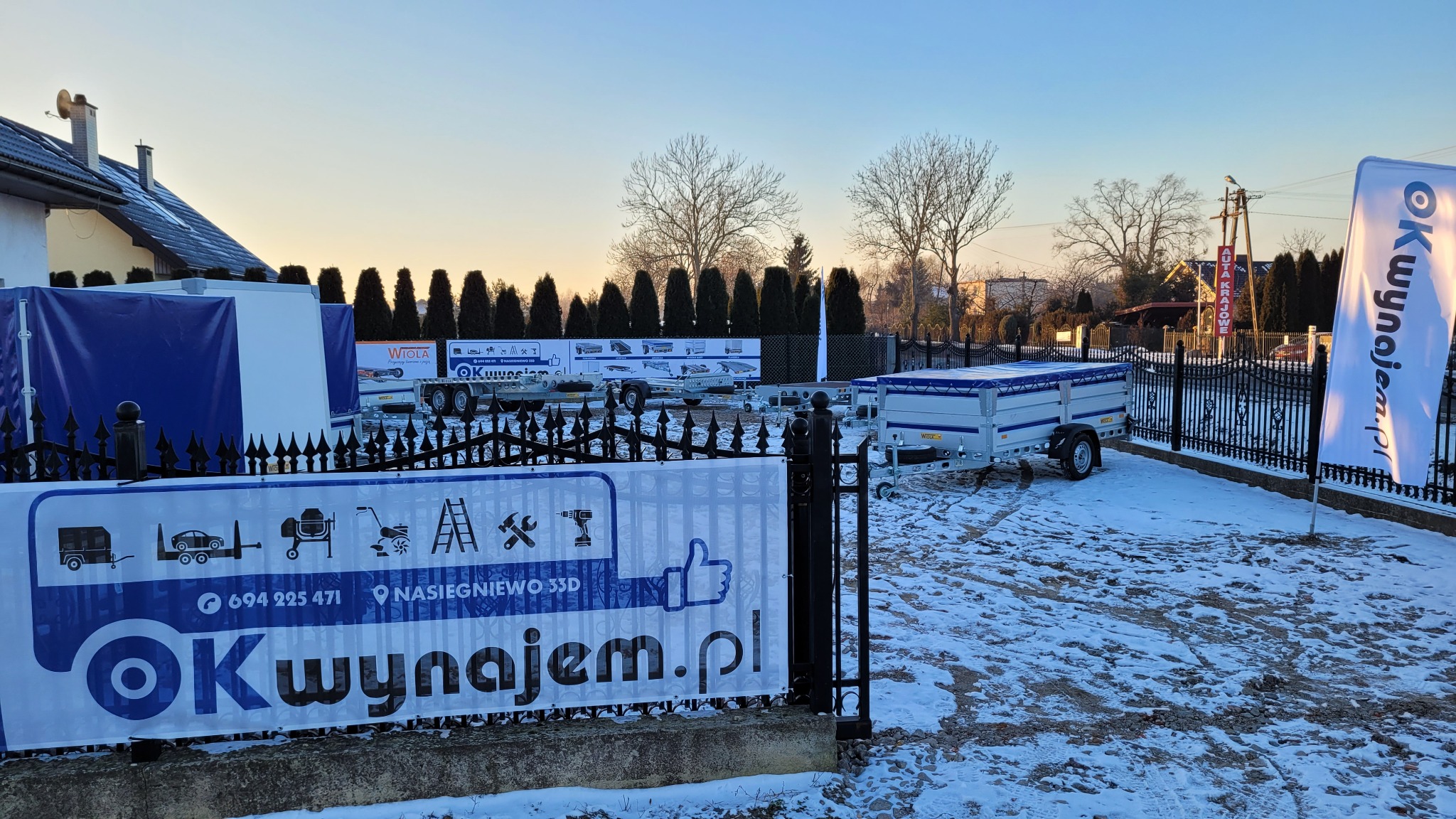 Plac z przyczepami do wynajęcia w zimowej scenerii, ogrodzony czarnym metalowym płotem, z banerem reklamowym i flagą firmy Okwynajem.pl.