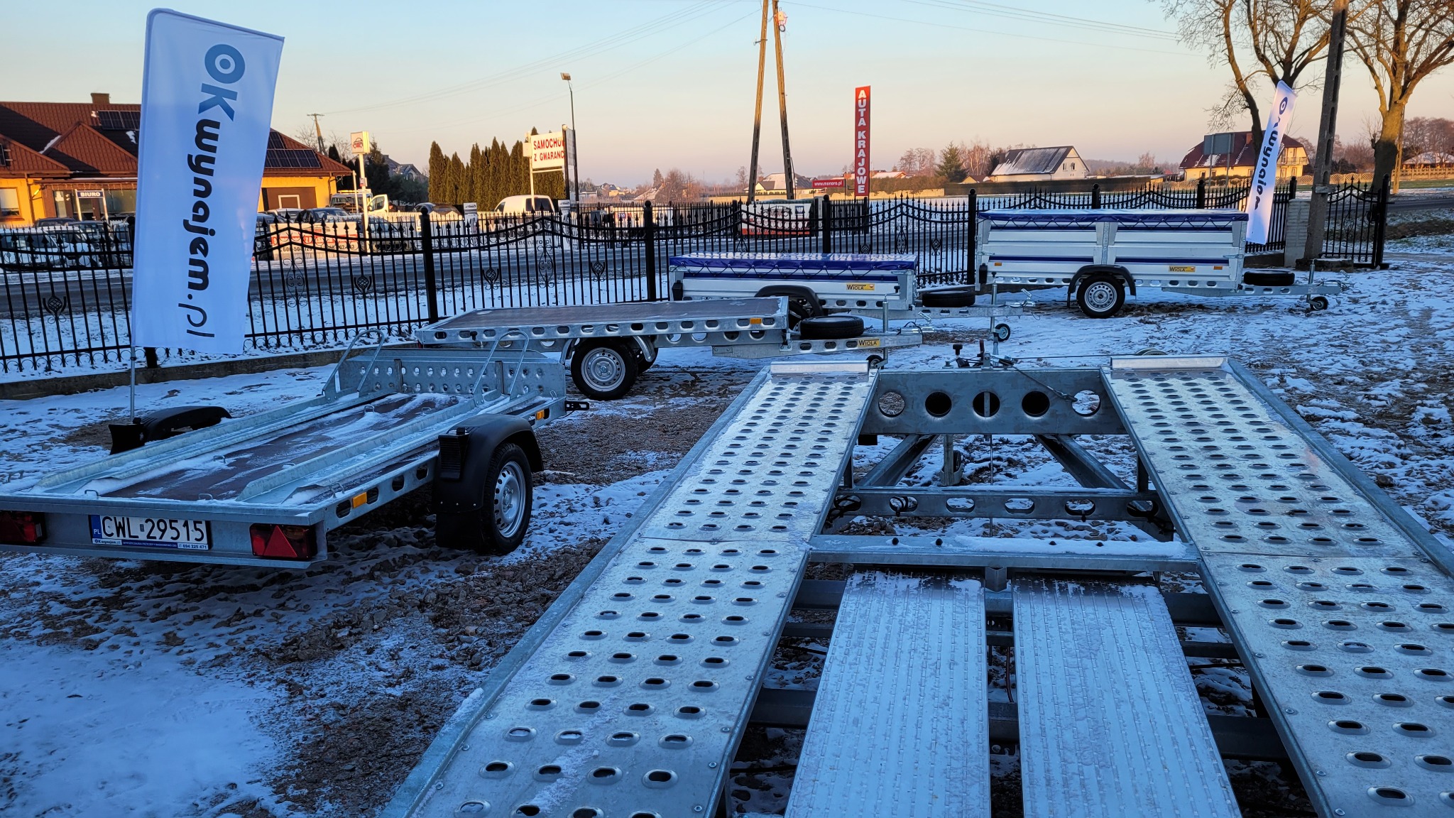 Różne typy przyczep samochodowych, w tym lawety, prezentowane na placu w zimowej scenerii, z widocznym logo firmy na fladze w tle.